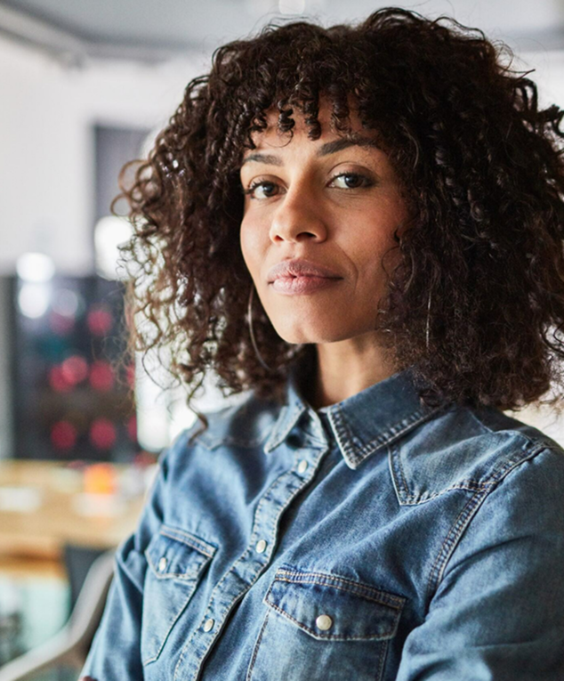 Jeune femme métis aux cheveux frisés et chemise en jean, regardant le visiteur.